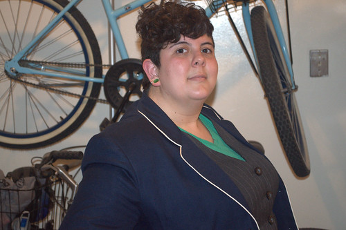 fat girl with short, asymmetrical curly hairdo in a green shirt, pinstripe vest, navy jacket with white piping, brown and rust striped skirt, green socks and houndstooth converse with green laces. Her finger nails are half houndstooth with a green stripe at the bottom of the pattern.