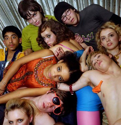 A group of nine teenagers lay on top of each other in a messy pile. They wear brightly colored clothes, except for one blonde boy toward the bottom right, who is shirtless. They are in front of a rippled silver backdrop.