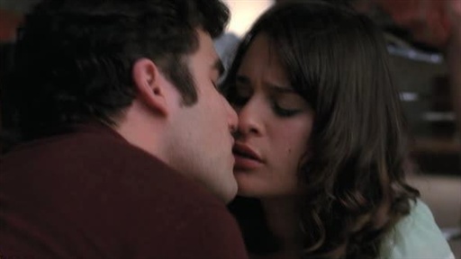 A man with dark curly hair and a dark red shirt leans in to kiss a woman with wavy brown hair. Their noses are touching and their eyes are closed.