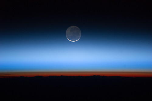 照片由國際太空站拍攝，月球在圖片中央，地球則靠近底部，紅橘色部份是對流層。照片由美國航太總署（NASA）提供。