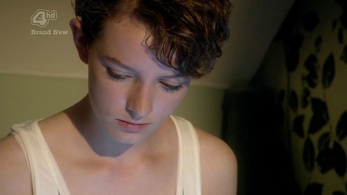 An adolescent girl with cropped curly hair looks downward. She is wearing a white tank top, and the shot is very tight on her face and upper torso. To the right of the frame is a wall with black decorative wallpaper. The camera is at an angle, so the white ceiling is also visible.