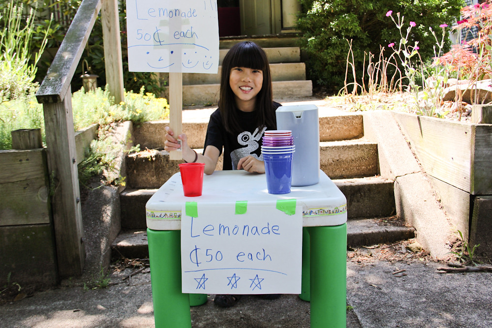 Young girl at the foot of stone steps in front of her house is sitting at a green and white table selling lemonade.
