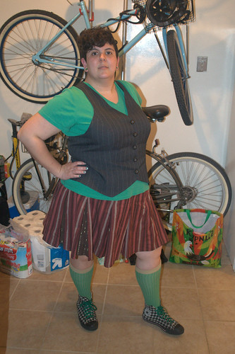 fat girl with short, asymmetrical curly hairdo in a green shirt, pinstripe vest, navy jacket with white piping, brown and rust striped skirt, green socks and houndstooth converse with green laces. Her finger nails are half houndstooth with a green stripe at the bottom of the pattern.