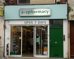 A small shopfront in green and white. A sign above reads “a-z pharmacy”, all in lower case with no spaces. The shopfront is fully glazed, and the door is open. Products on shelves are visible through the glass.