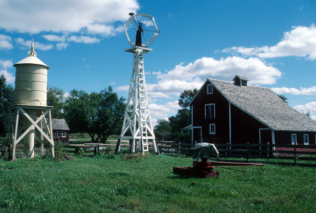 An old farmhouse, windmill, and water tower on the grounds of the Stuhr Museum of the Prairie Pioneer in Nebraska. The buildings stand against a picture-perfect blue and white cloudy sky.